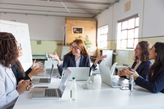 Cheerful businesswomen working together in office. Multiethnic female colleagues sitting around table with laptops and discussing work in office