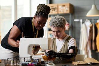 Two young female specialists in sewing clothes working together in workshop one amputee