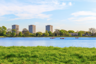 Panoramic view of a calm lake with three orange boats, lush green grass in the foreground, and several tall apartment buildings and trees on the opposite shore under a blue sky.