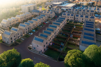 Aerial view of a suburban neighborhood showcasing rows of brick-clad houses with solar panels installed on their rooftops. The houses are arranged along winding streets and have small backyards. Trees with green foliage are present in the foreground. In the background, there's an apartment building and other buildings.