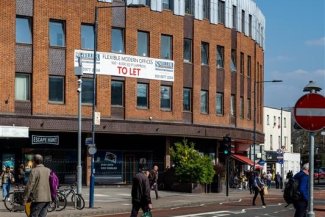 A group of people crossing the street in front of a large vacant commercial space on a busy urban street.