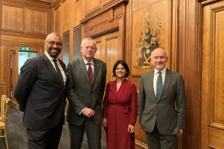 Rt Hon Sir James Cleverly, Peter Hogg, Muniya Barua, and John Dickie in formal attire ,standing before a wooden panel.