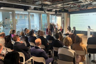 A diverse group of people in chairs, engaged in a discussion while facing a large screen in a conference setting.
