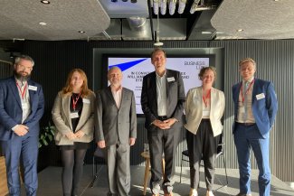 Six individuals in business attire, including John Dickie of BusinessLDN and Alex Williams of TfL smile to camera in front of screen at an event 