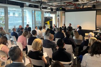 A group of people attend a business panel discussion in a modern conference room, seated and facing speakers at the front with a screen and "BusinessLDN" banner.