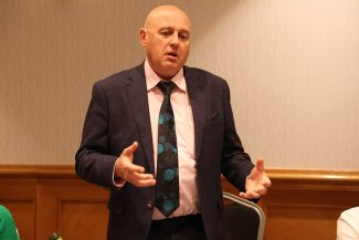 A man in a suit and tie stands at a podium, delivering a speech to an audience.