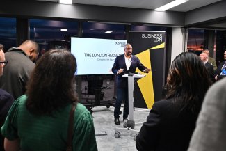 James Cleverly MP addresses a packed room at a reception. Screen behind him bears legend - London Reception BusinessLDN